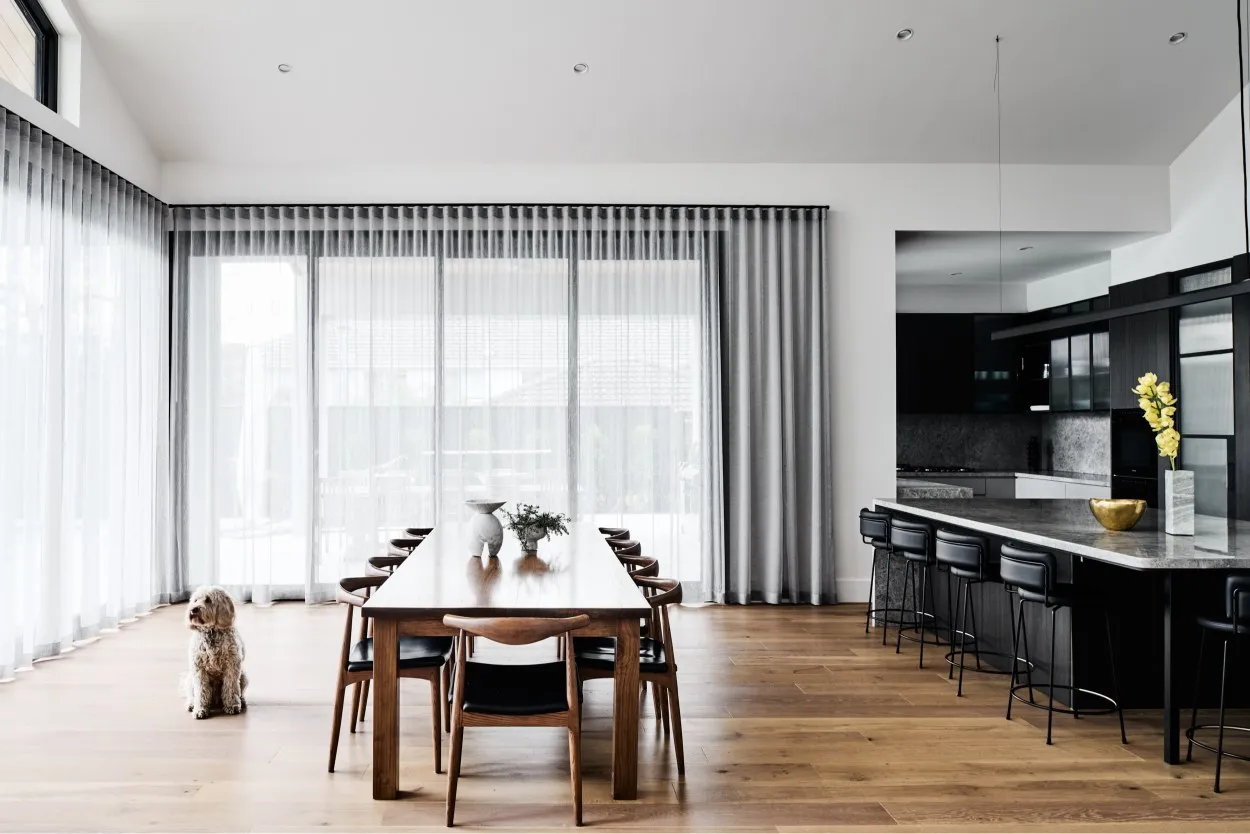 Open-plan dining room with long timber table, sheer curtains, and dark kitchen beyond