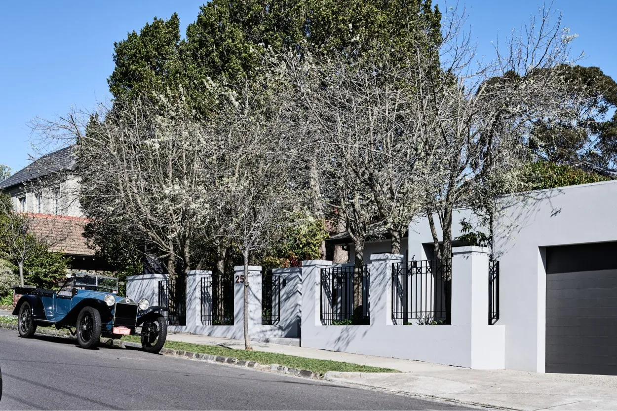 Street-facing rendered home exterior with black iron fence, mature trees and vintage car parked outside