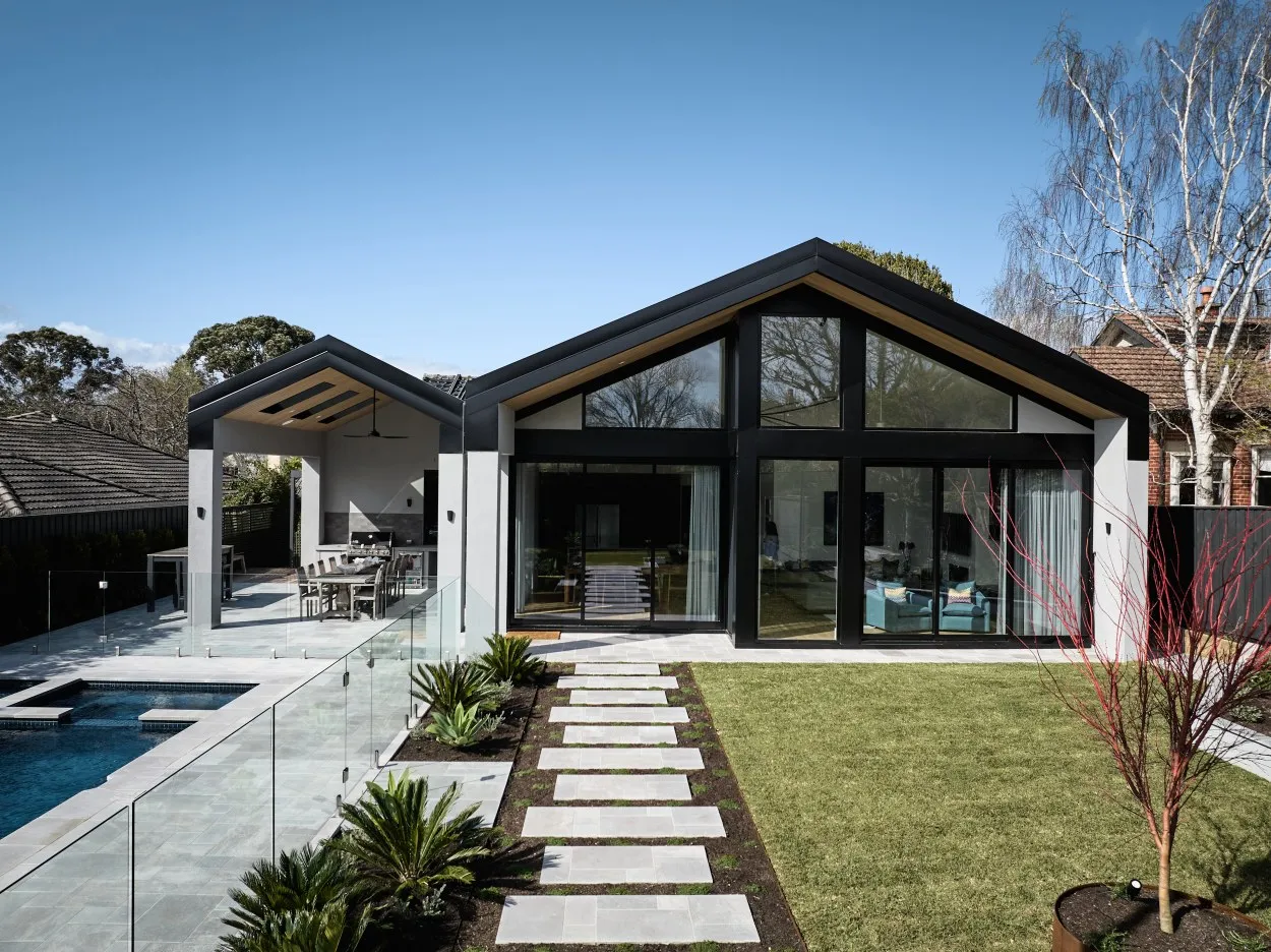 Double-gable home exterior viewed from rear garden with pool, stepped path and floor-to-ceiling glazing
