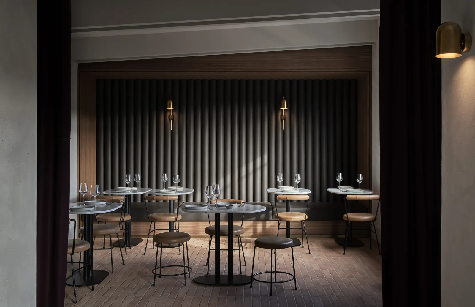 Intimate restaurant dining room with fluted grey wall panels, brass wall sconces and round marble-top tables