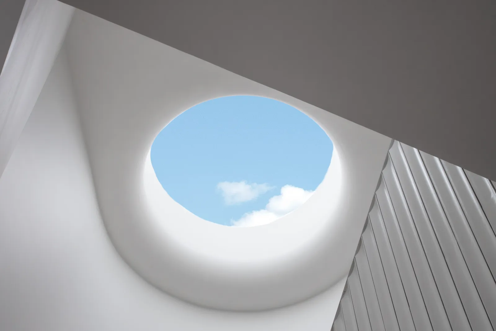Upward view of circular skylight framing blue sky with ribbed white wall panelling alongside