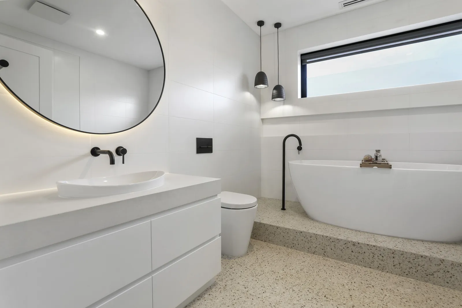 Bathroom with freestanding bath on a terrazzo platform, round mirror and letterbox window