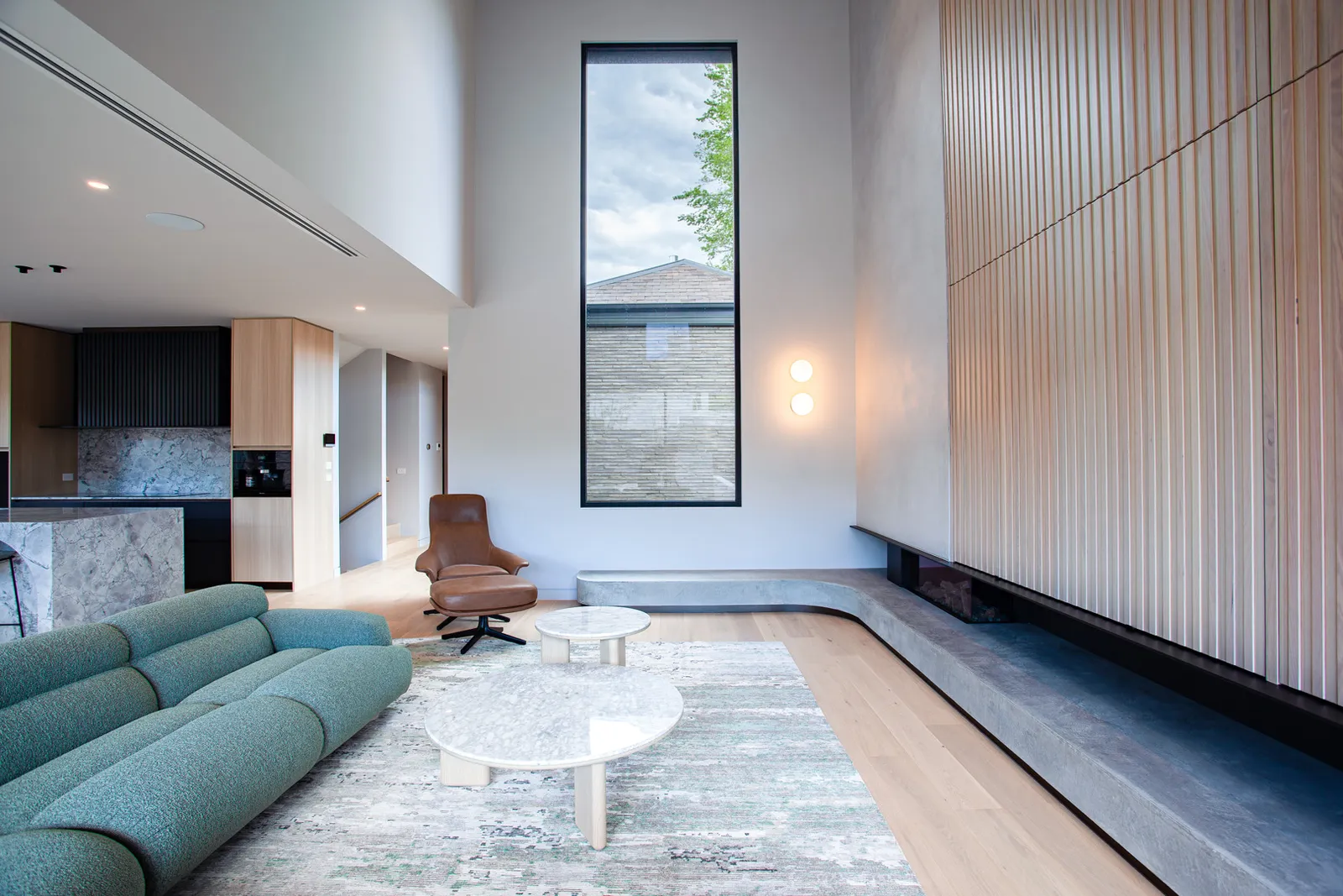 Double-height open-plan living room with teal sofa, timber slat wall, tall window and kitchen beyond