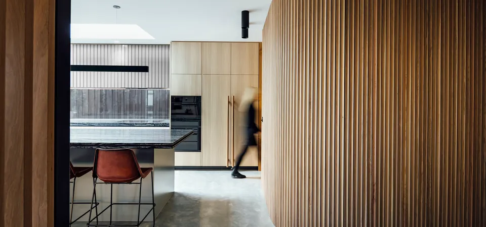 Curved timber batten column beside dark stone kitchen island with leather barstools and concrete floor