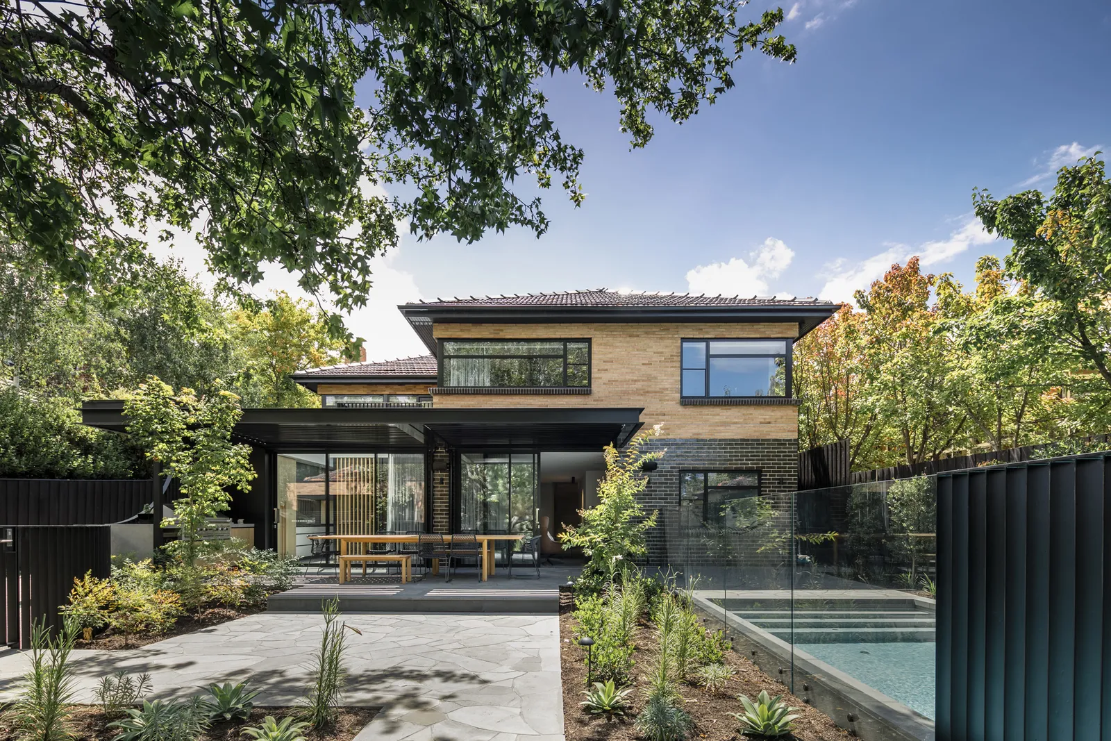 Two-storey brick and dark timber home exterior viewed from garden with pool and patio