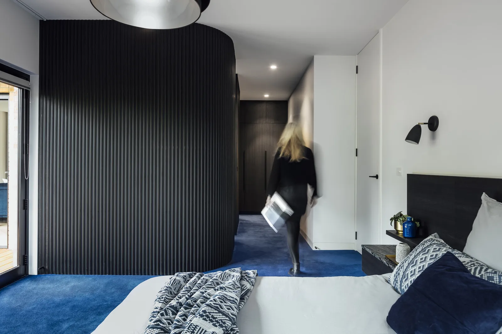 Contemporary bedroom with dark fluted curved wardrobe, blue carpet and figure in motion