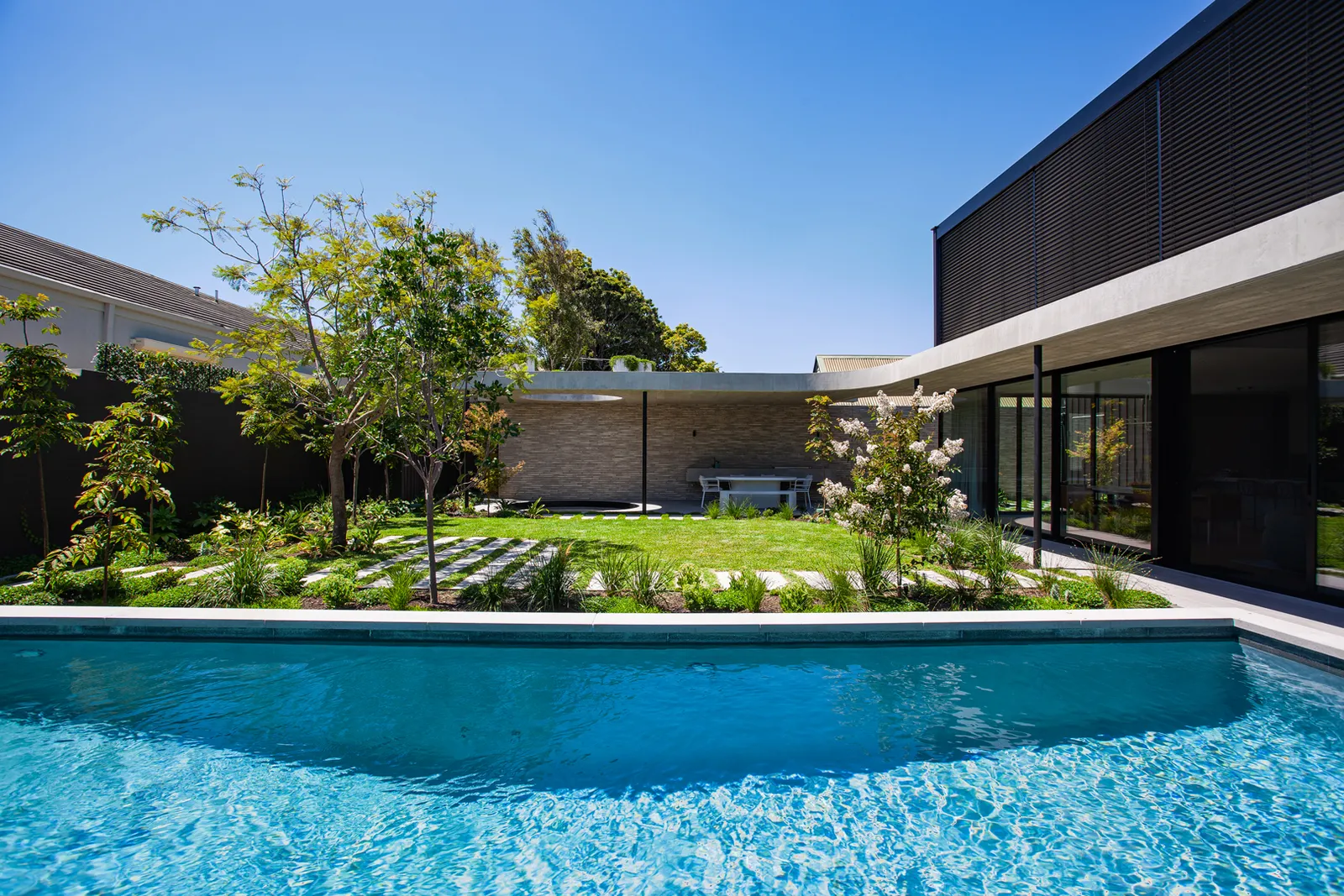 Outdoor pool with lush garden, stepping stones, and modern black-clad residence under blue sky