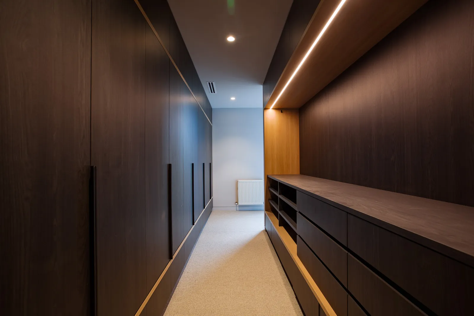 Long walk-in wardrobe with dark timber joinery, LED strip lighting and built-in drawers
