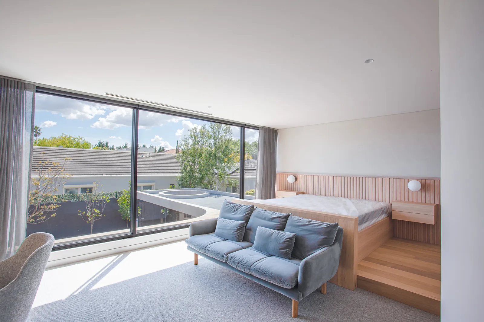 Bright master bedroom with elevated timber bed platform, blue sofa, and panoramic floor-to-ceiling windows