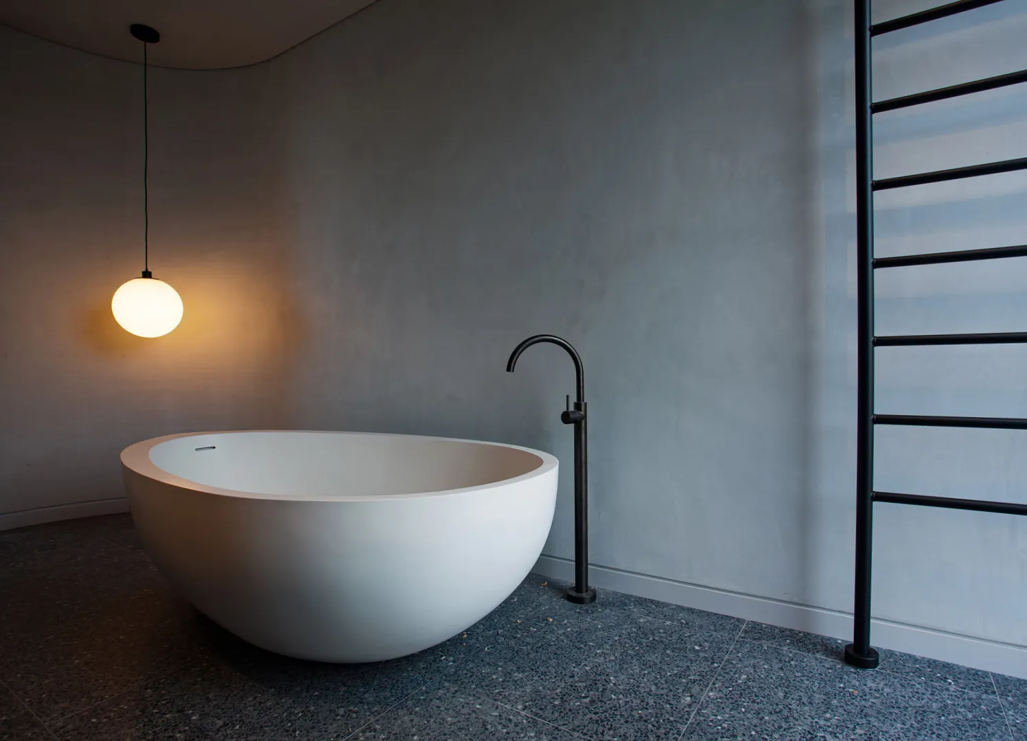 Freestanding white oval bath with black tapware, pendant light, and terrazzo floor