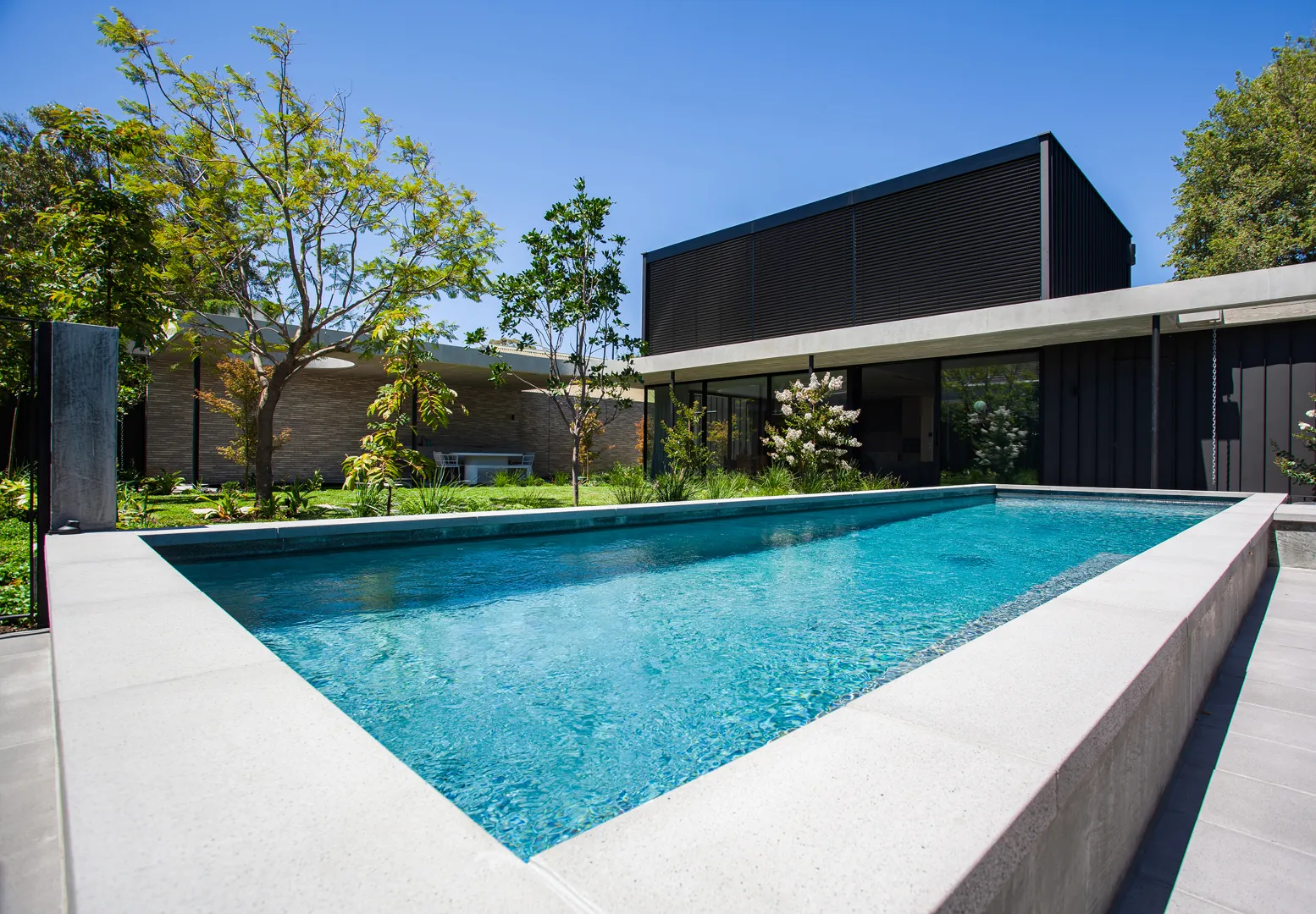 Rectangular lap pool with blue water beside a modern dark-clad two-storey residence surrounded by mature gardens