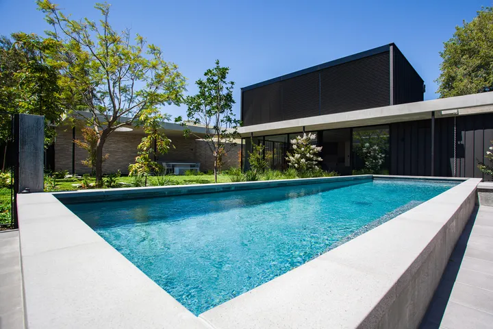 Rectangular lap pool with blue water beside a modern dark-clad two-storey residence surrounded by mature gardens