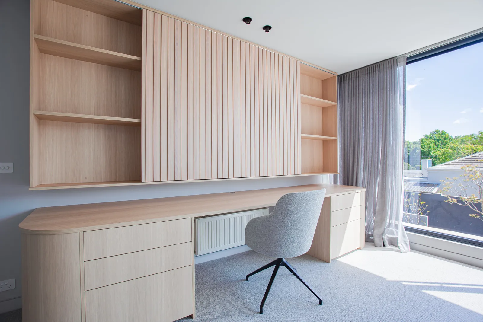 Timber built-in study desk with open shelving, slatted panels, and large window