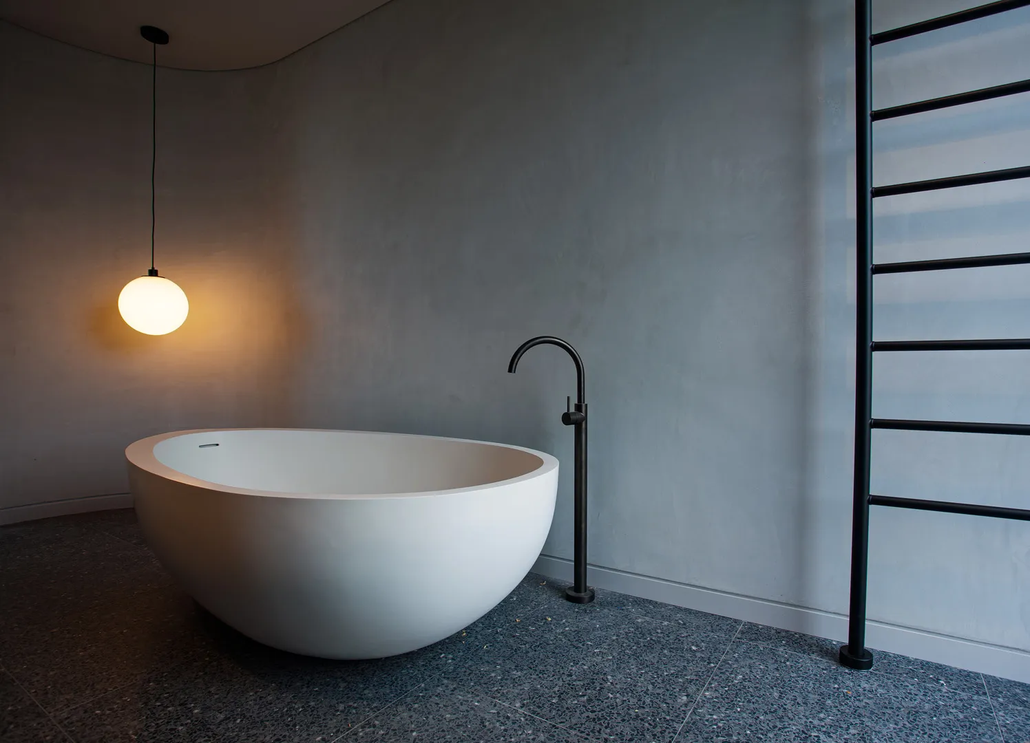 Freestanding oval bathtub with matte black floor-mounted tap, pendant light and concrete-look walls