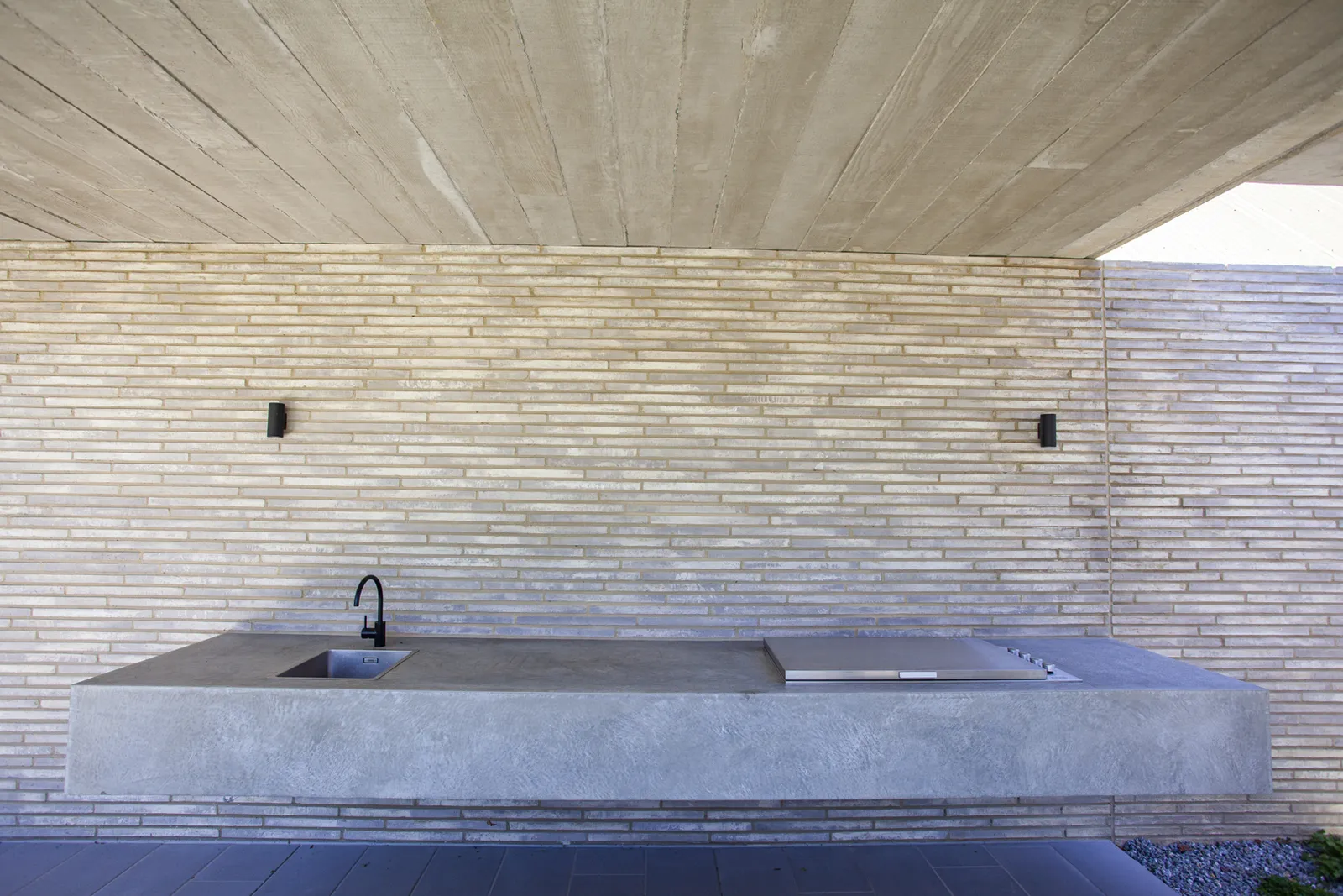 Outdoor kitchen bench in concrete with black tap set against textured brick wall under concrete ceiling