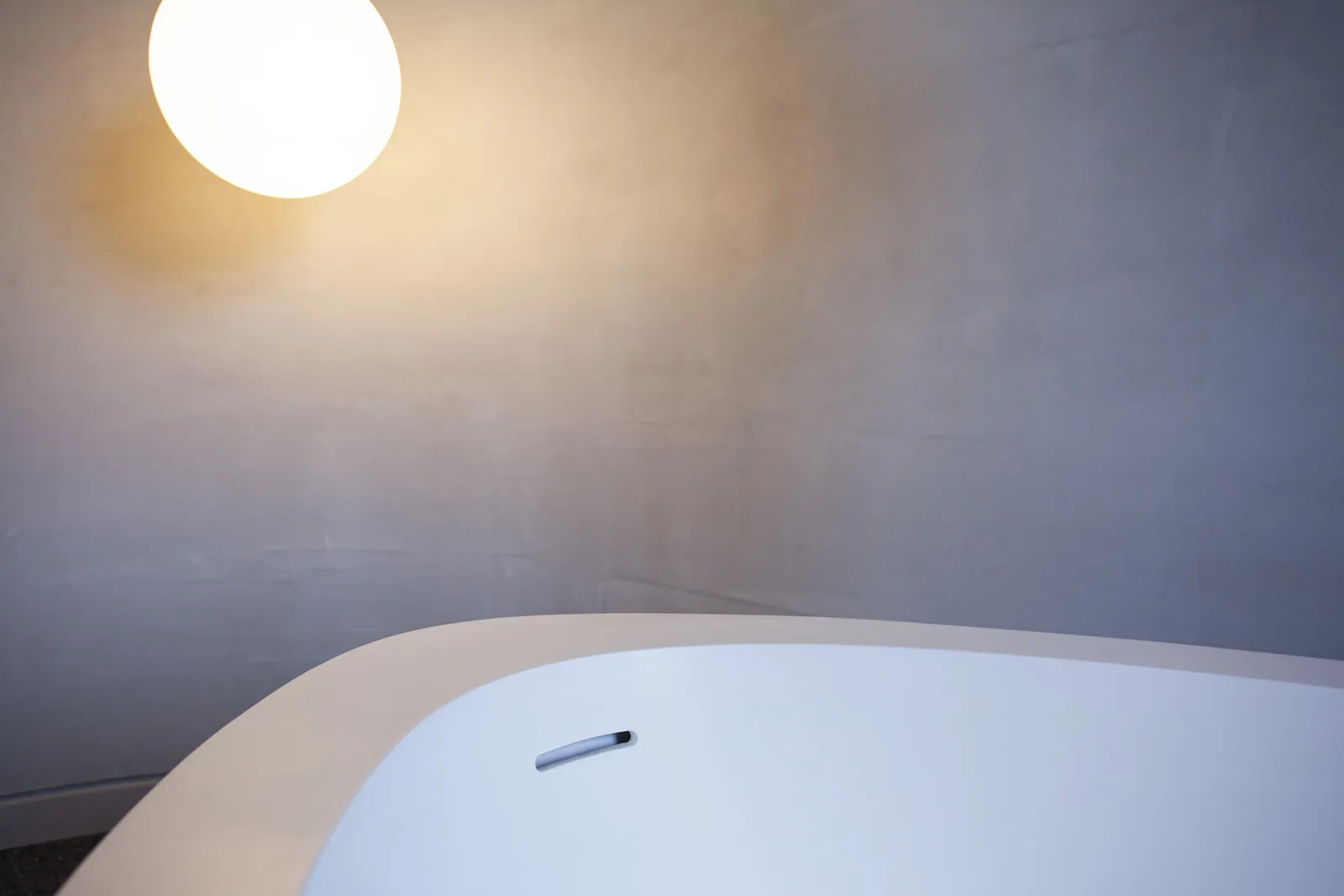 Freestanding white bathtub beneath a glowing globe pendant light in a minimal plaster bathroom