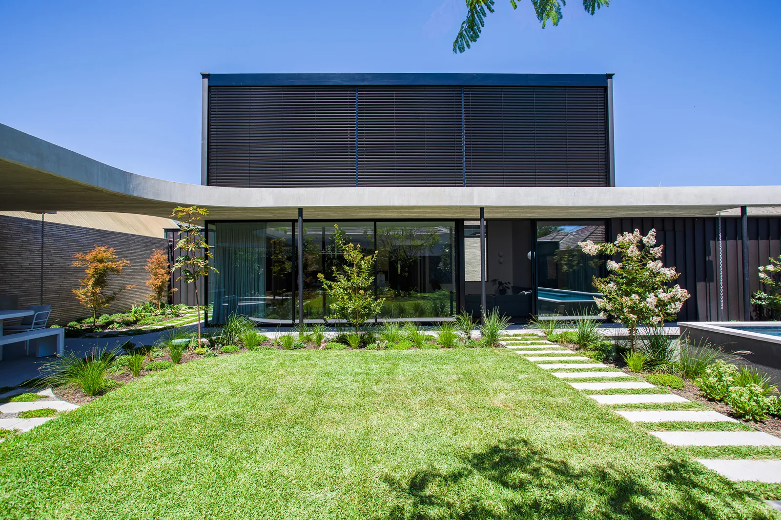 Rear garden view of a contemporary dark-clad home with floor-to-ceiling glazing, stepping stone path, and lawn