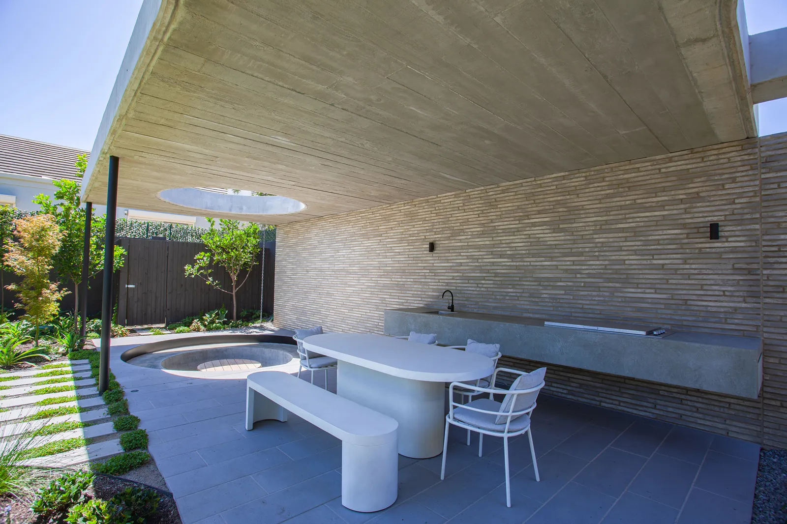 Covered outdoor dining area with concrete ceiling, brick wall, white table and bench beside circular sunken garden