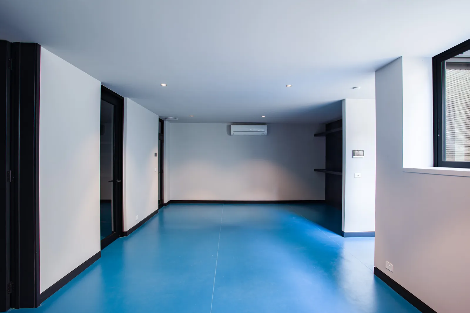 Empty lower-level room with bold blue epoxy floor, split-system air conditioner, and black-framed windows