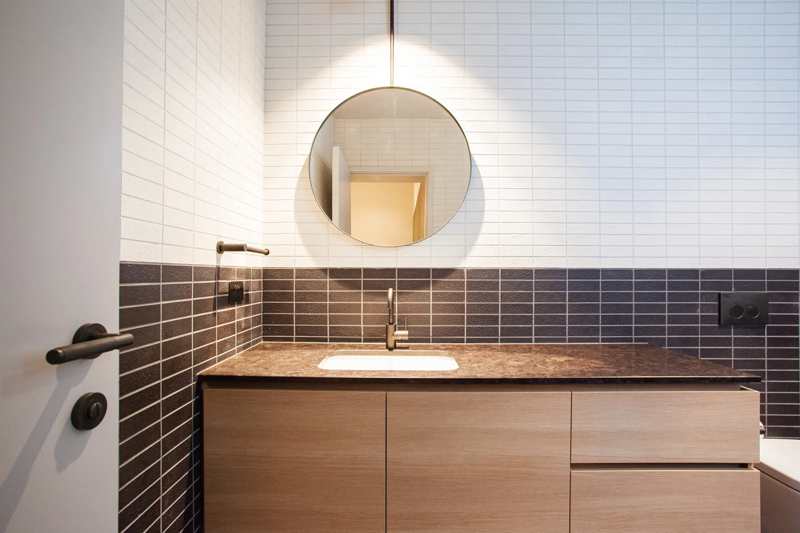 Bathroom vanity with round mirror, two-tone tile splashback, stone benchtop and timber cabinetry