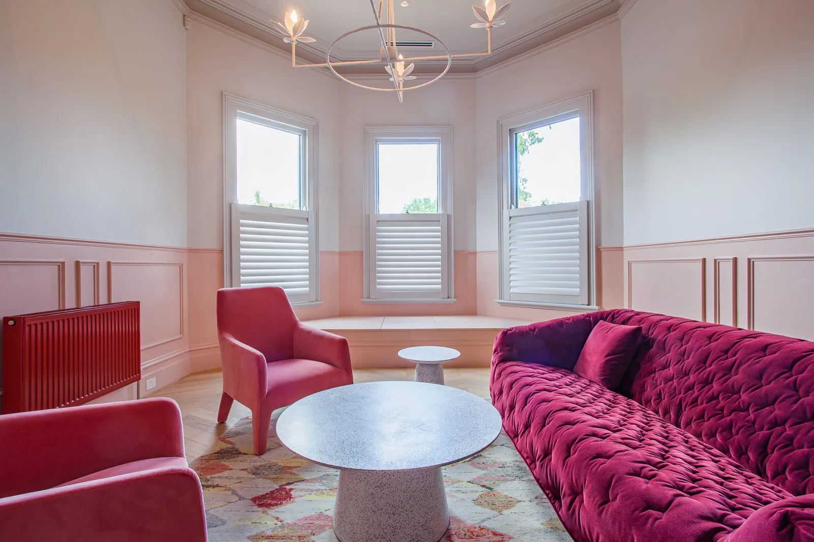 Bay window sitting room with tufted magenta sofa, pink armchairs, wainscoting, and floral chandelier