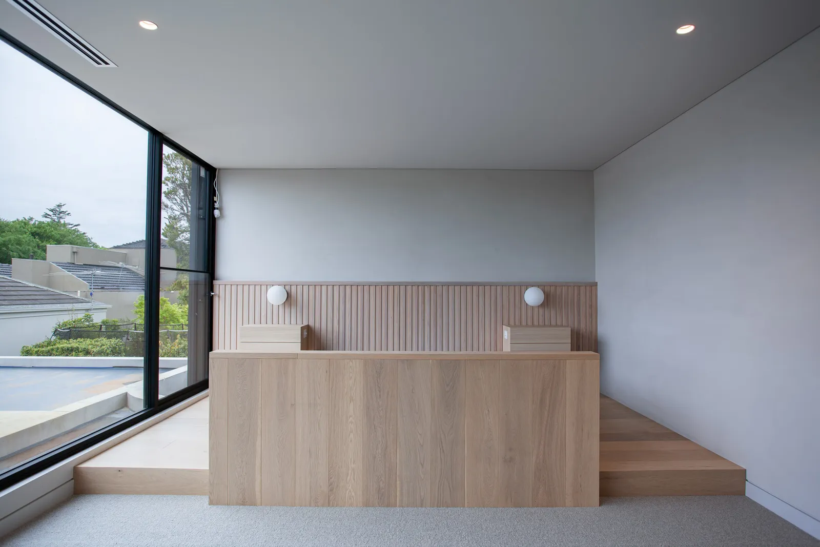 Elevated timber bed platform with oak headboard, globe wall sconces, and floor-to-ceiling window