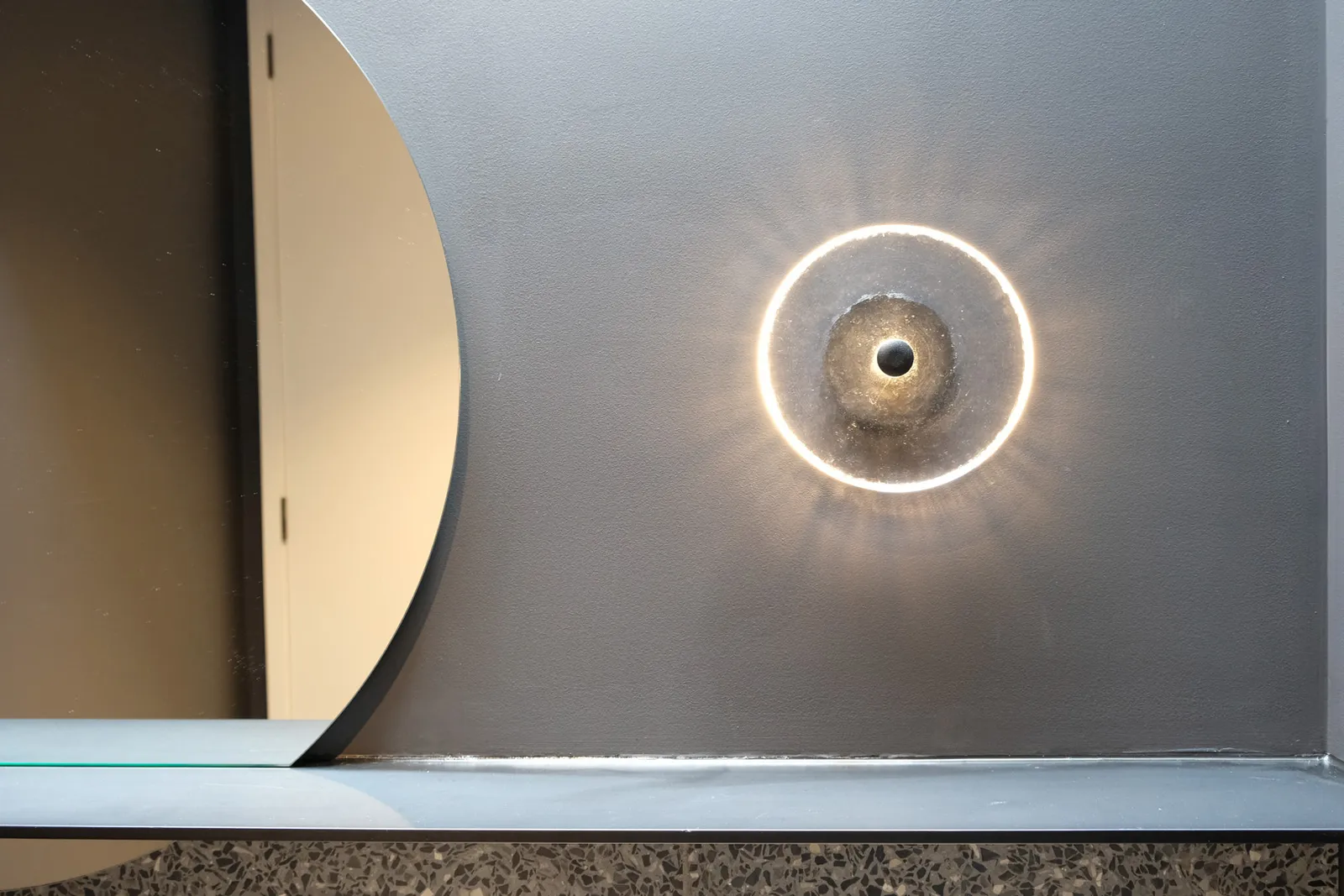 Circular LED wall light casting halo glow on grey wall beside round mirror and stone vanity