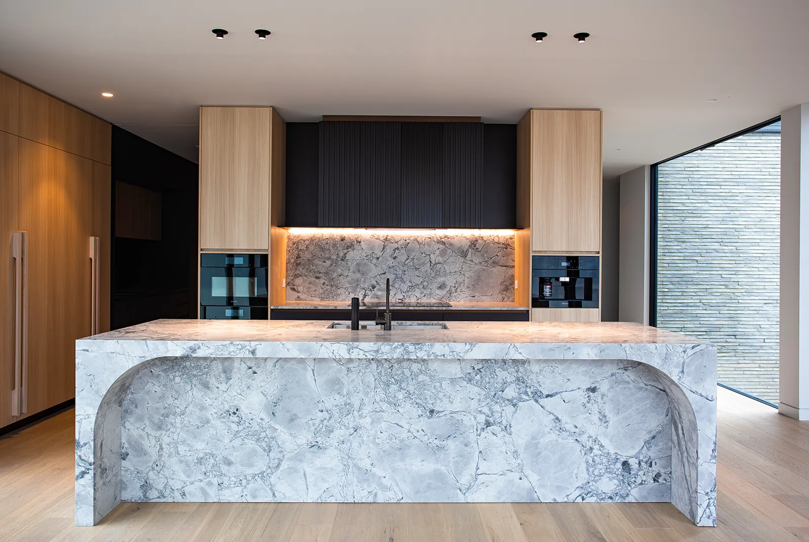 Open-plan kitchen with curved marble island, timber cabinetry, black tapware, and floor-to-ceiling glazing
