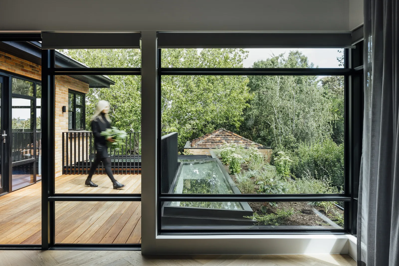 Person walking on timber deck viewed through large black-framed windows overlooking garden