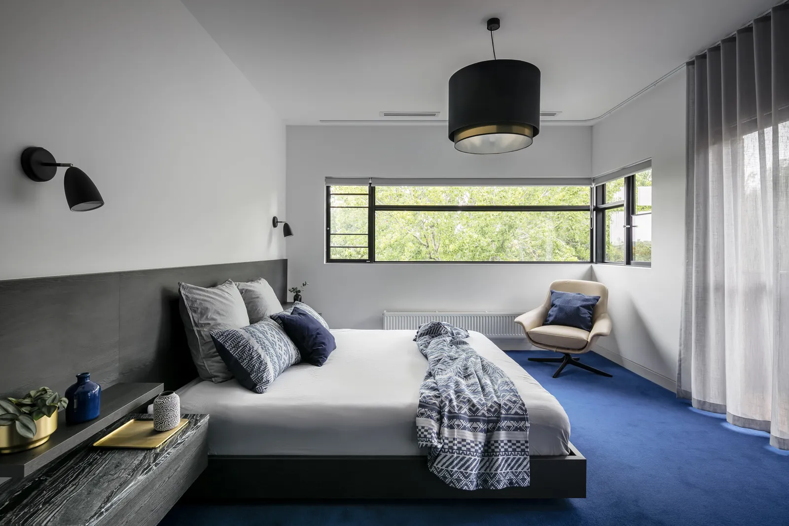 Master bedroom with blue carpet, low platform bed, black pendant light, and garden window