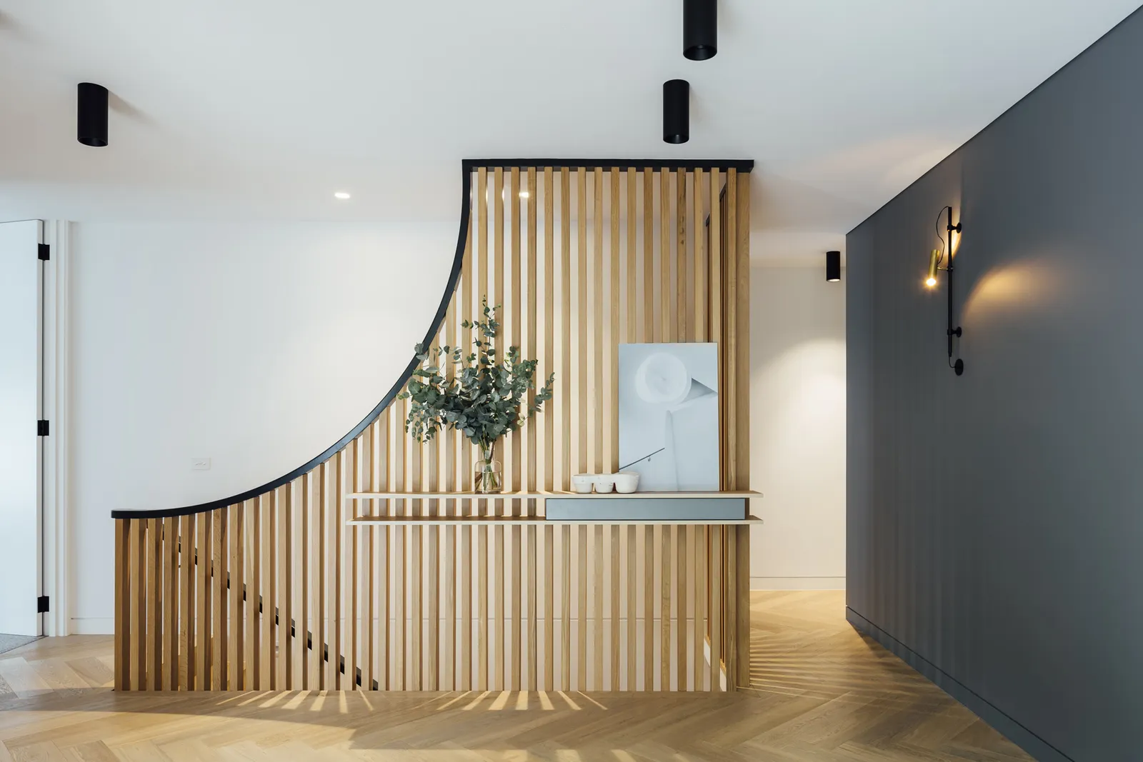 Curved timber batten staircase balustrade with console shelf, herringbone floor, and wall sconce