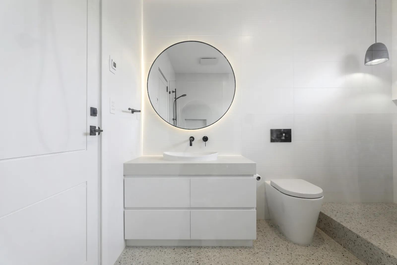 Modern white ensuite with round backlit mirror, vessel sink vanity, and terrazzo floor