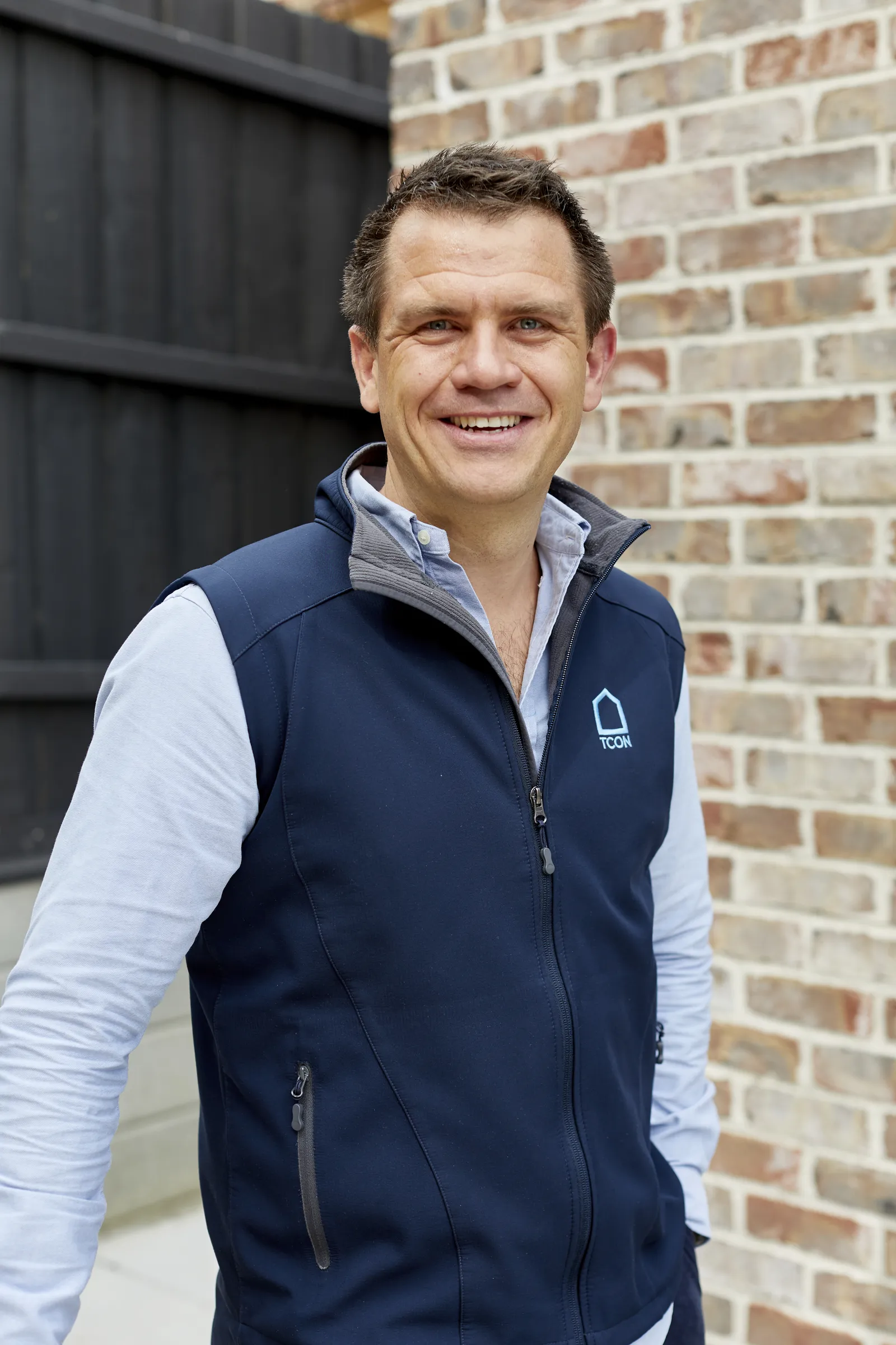Smiling man in TCON navy vest standing against a brick wall outdoors