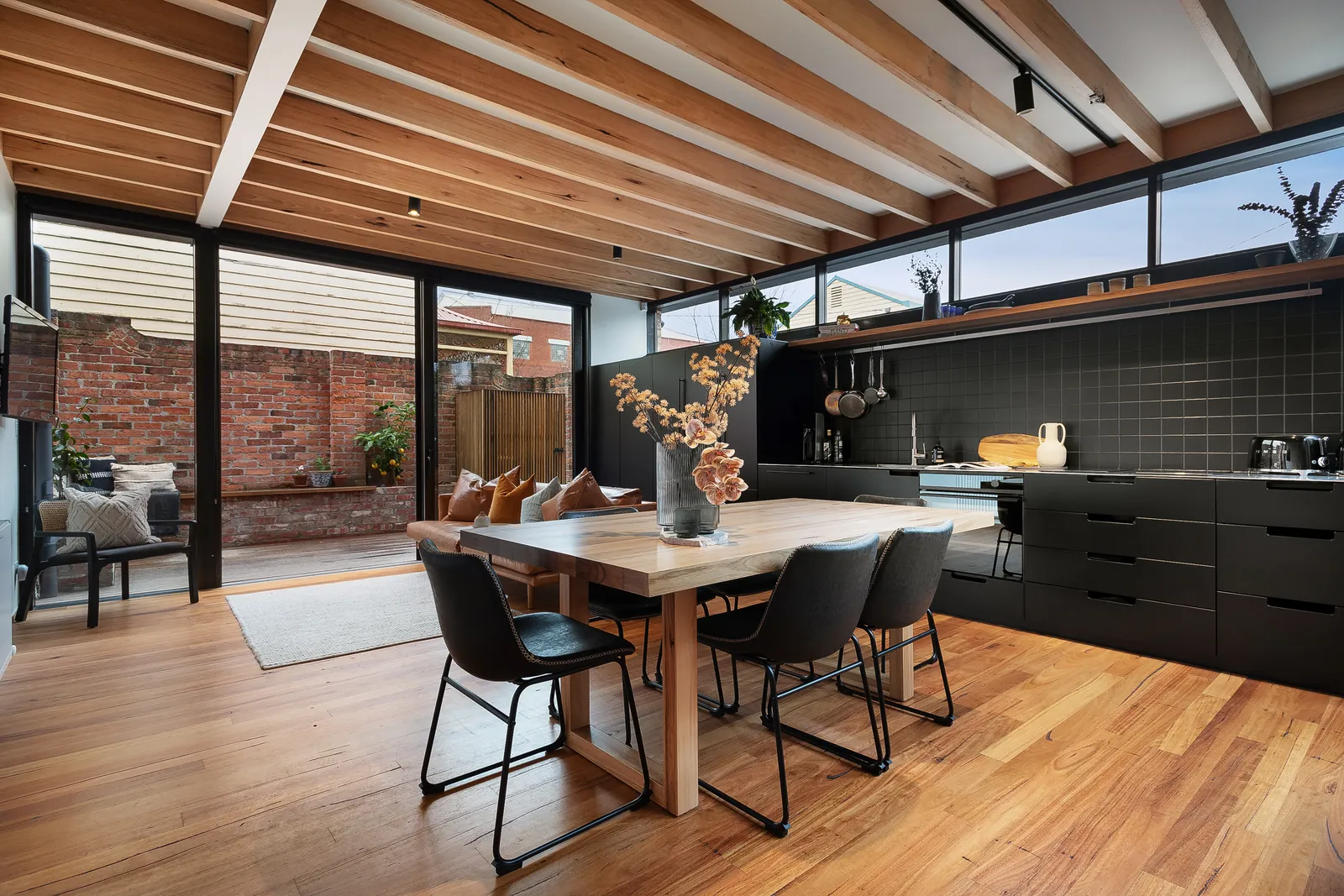 Dining area with timber table, black chairs, exposed timber beams, dark kitchen, and courtyard view