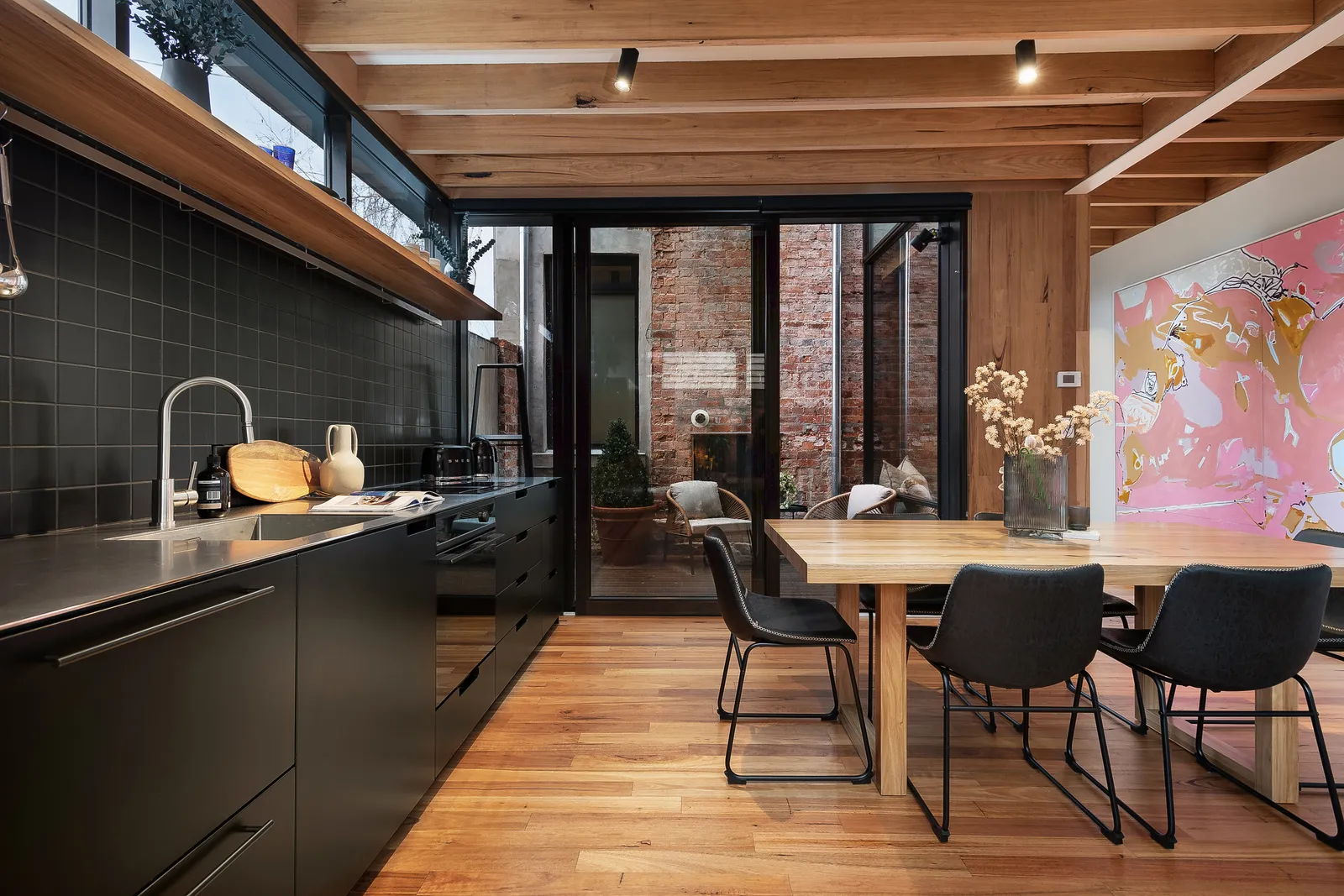 Open-plan kitchen and dining with black cabinetry, timber ceiling beams and courtyard outlook