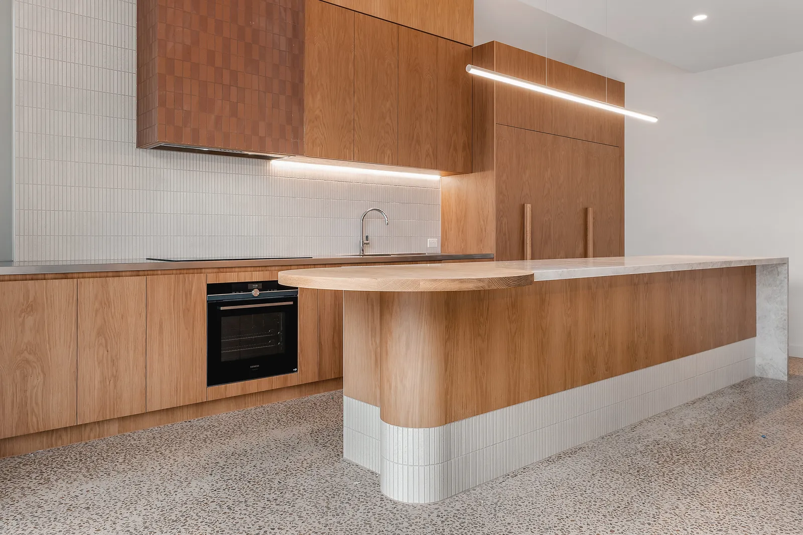 Open-plan timber kitchen with island bench, black oven, white tile splashback and terrazzo floor