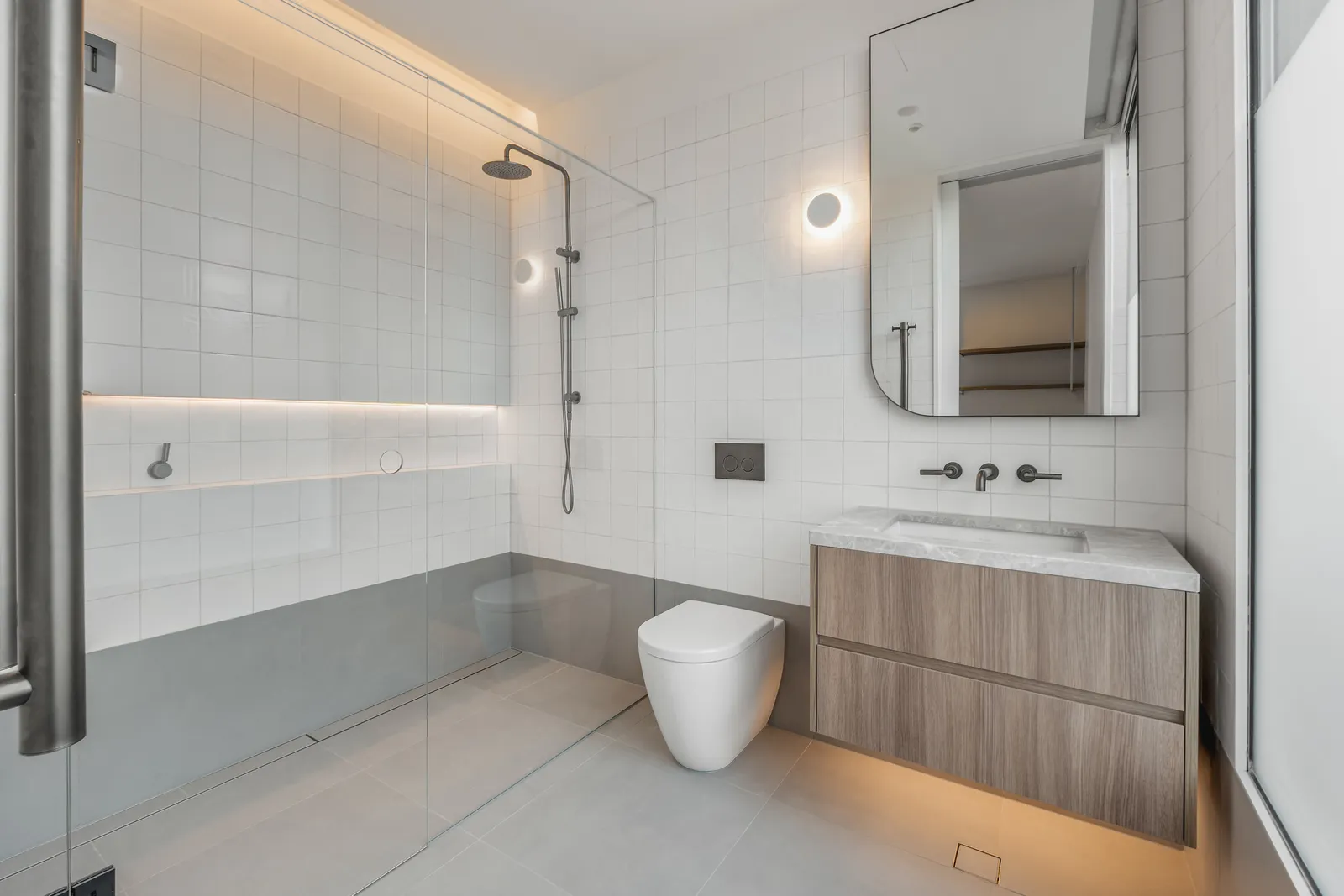 Light-filled bathroom with frameless glass shower, floating timber vanity, and round wall sconce