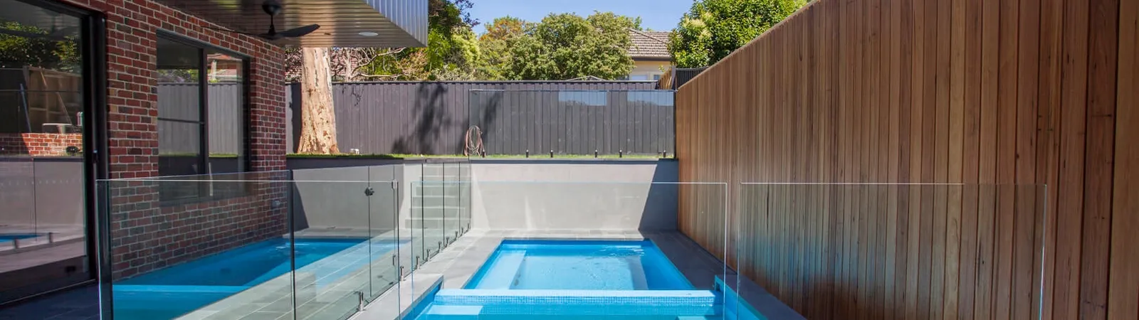 Outdoor pool and spa with glass fencing, timber screen wall, and blue sky