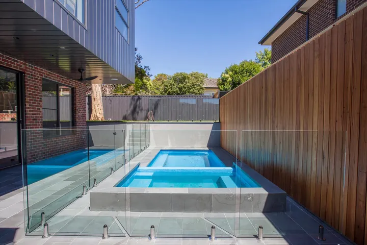 Outdoor pool and spa with glass fencing, timber screen wall, and blue sky