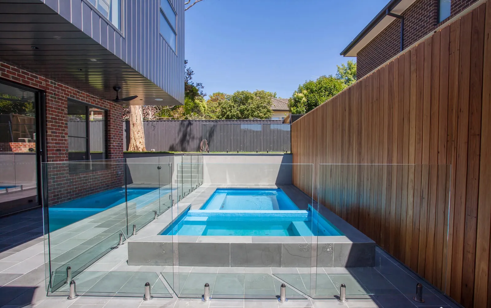 Outdoor pool and spa with glass fencing, timber screen wall, and blue sky