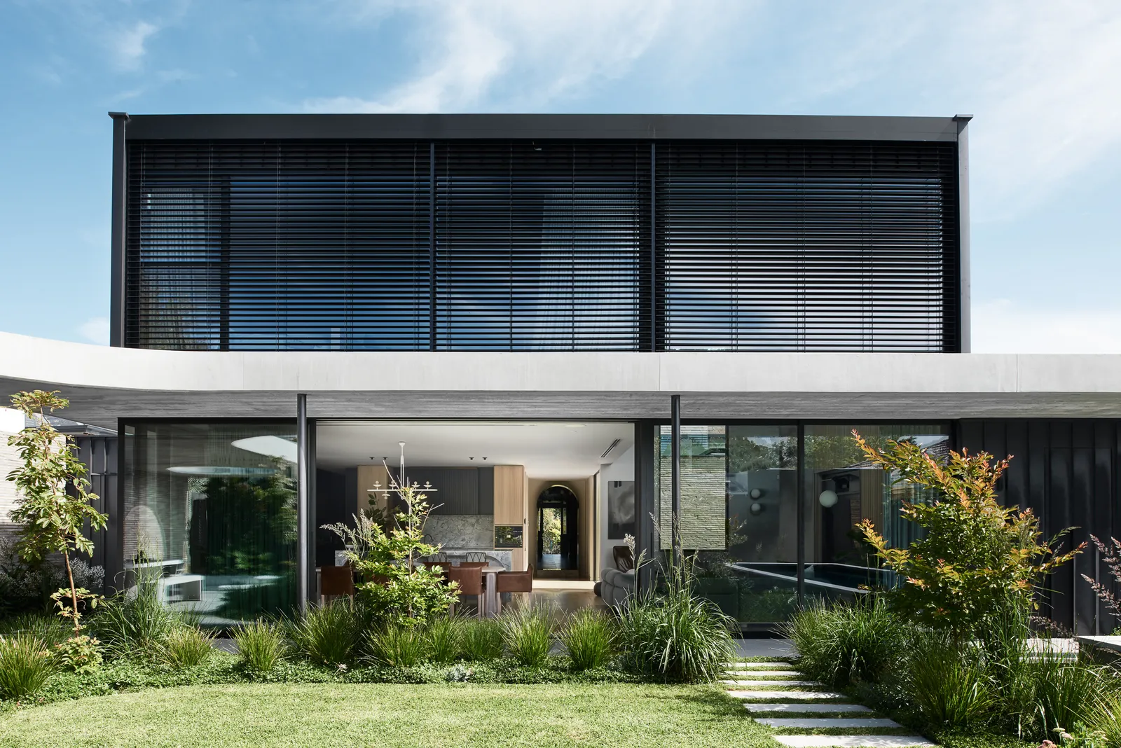 Modern two-storey home facade with dark louvres, glazed ground floor, lawn and native plantings