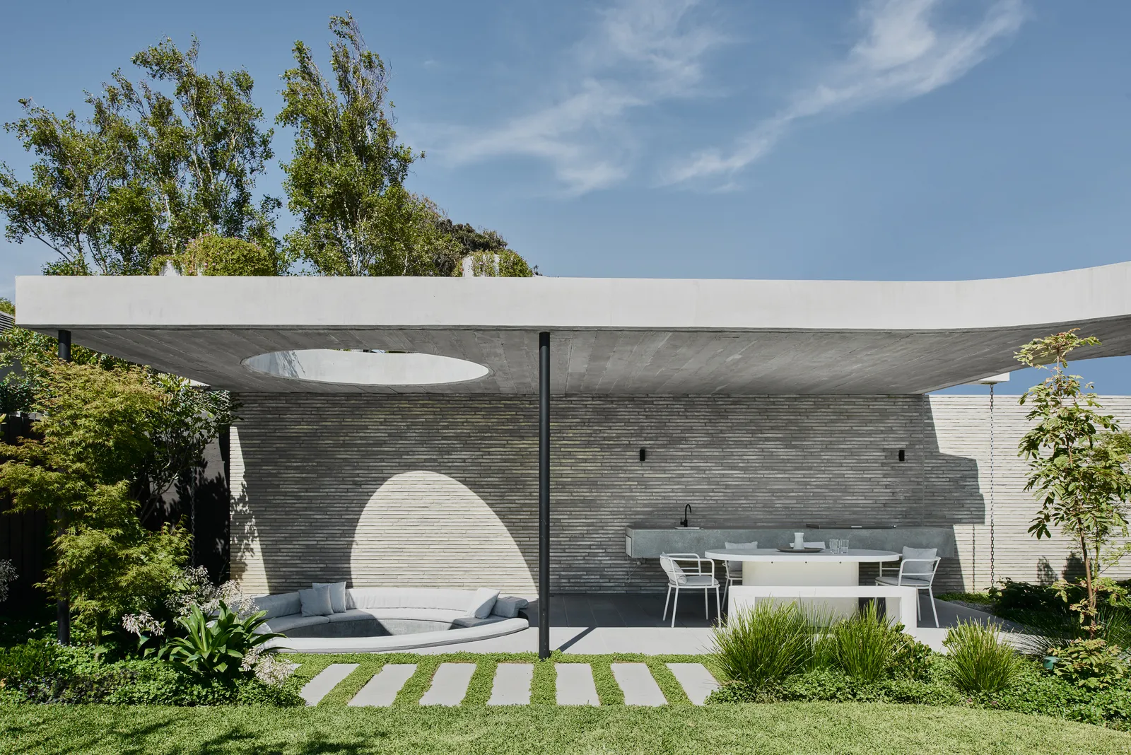 Concrete outdoor pavilion with circular skylight cutout, built-in bench, outdoor kitchen, and manicured lawn