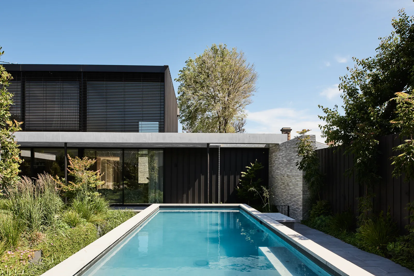 Rear exterior of dark-clad two-storey home with lap pool, lush garden and louvred upper facade