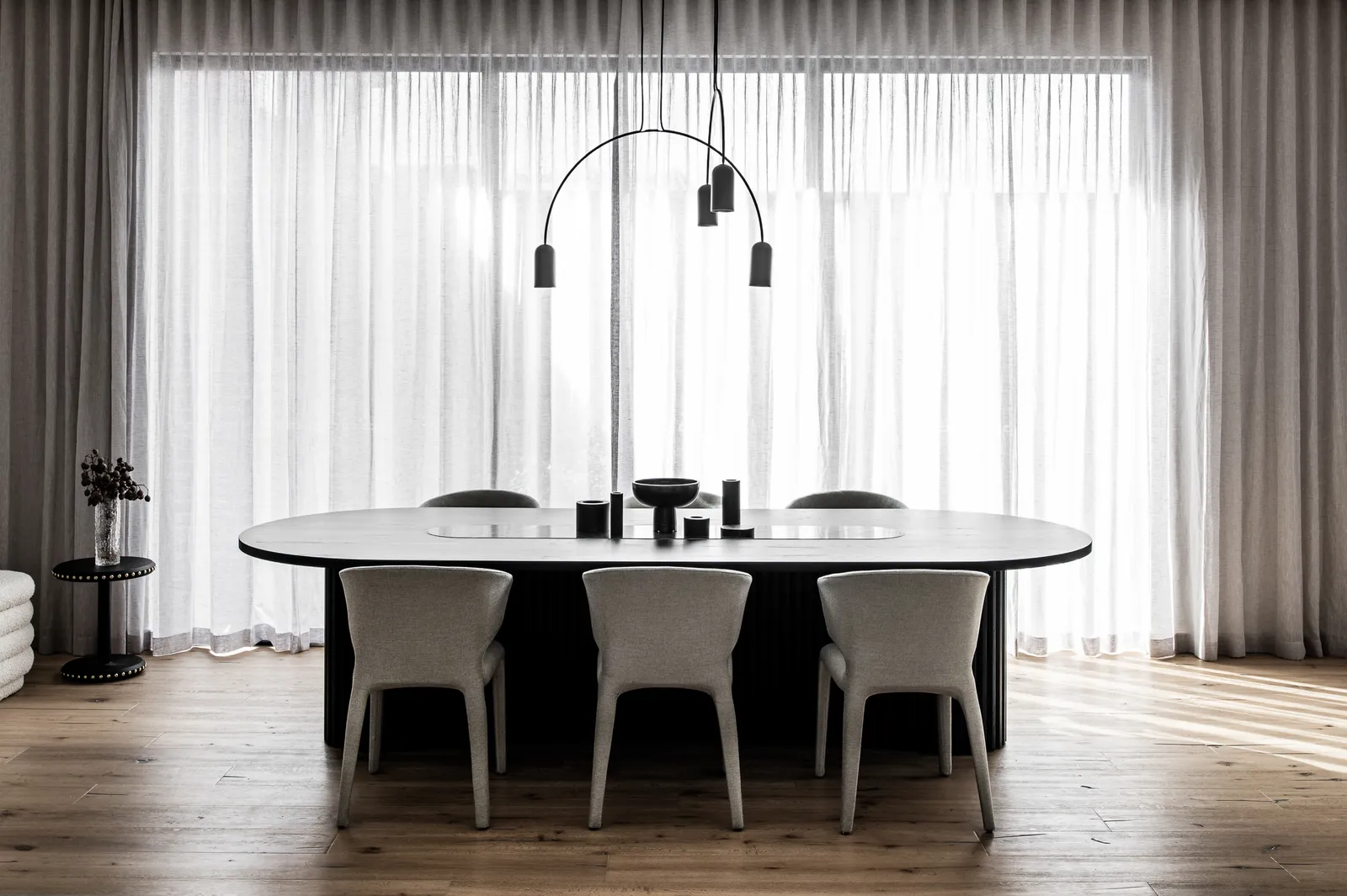 Minimalist dining room with oval table, upholstered chairs, and arc pendant over sheer curtains