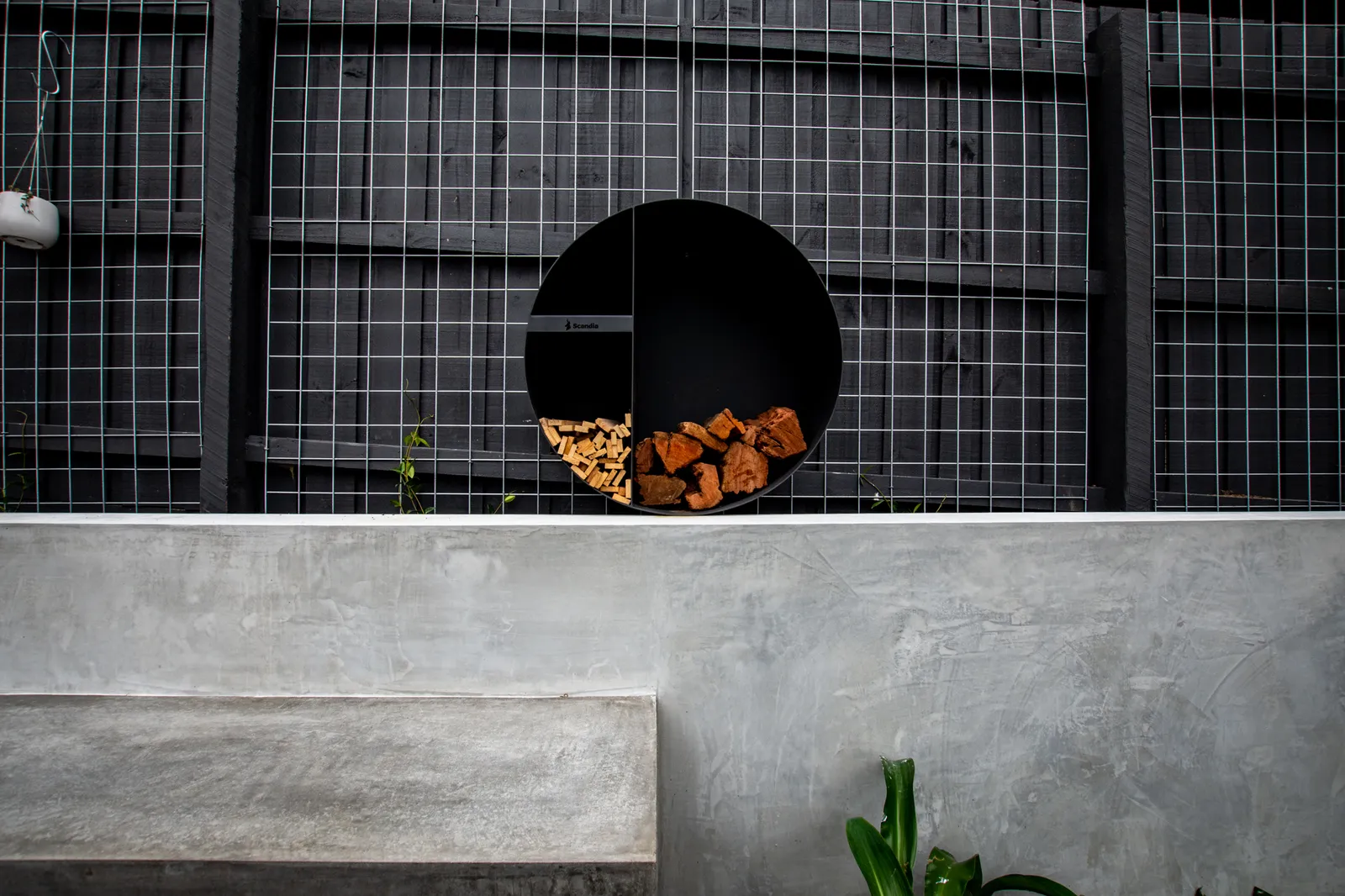 Circular black outdoor fireplace with firewood storage against wire mesh fence and concrete bench